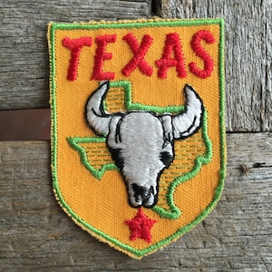 May include: A yellow felt patch with green stitching, depicting the outline of the state of Texas with a white skull and crossbones inside. The word "TEXAS" is embroidered in red above the state outline.