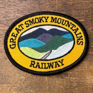 May include: An oval embroidered patch featuring a yellow border with the black text GREAT SMOKY MOUNTAINS RAILWAY. The center depicts a landscape of layered blue, green, and light green mountains with a white path leading toward the peaks.