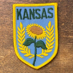 May include: A shield-shaped embroidered patch with a light blue background and a yellow border. The word KANSAS is stitched in dark green at the top. Below the text is a yellow sunflower with a brown center, flanked by two yellow wheat stalks.