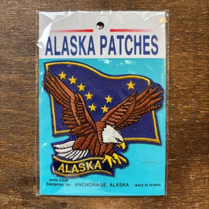 以下が含まれることがあります： 刺繍デザインが施された、パッケージ入りのアラスカパッチ。パッチは、金色の星が散りばめられた青い背景に、翼を広げたハクトウワシと、その下のバナーに「ALASKA」の文字が描かれています。「ALASKA PATCHES」と書かれたパッケージ。