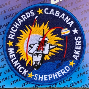 May include: Circular embroidered patch with a blue border, depicting a space shuttle, sun, and stars. The names "RICHARDS CABANA AKERS MELNICK SHEPHERD" are embroidered around the edge. The patch has a detailed design with red, yellow, and white accents, and is likely a collectible item.