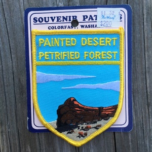 Puede incluir: Un parche bordado amarillo y azul con el texto "Painted Desert Petrified Forest" y una representación de una formación rocosa roja sobre un fondo de cielo azul.