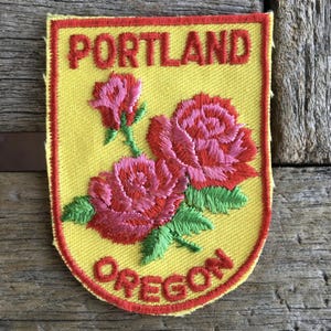 Könnte beinhalten: Ein schildförmiger, bestickter Aufnäher mit gelbem Hintergrund und rotem Rand. Der Text PORTLAND ist oben in Rot gestickt, und OREGON ist unten in Rot gestickt. Zwei rosa und rote Rosen mit grünen Blättern befinden sich in der Mitte.