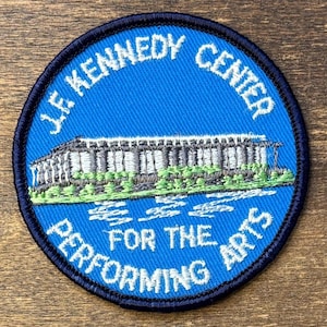 May include: A circular embroidered patch with a bright blue background and a dark blue border. It features a white and grey stitched illustration of the J.F. Kennedy Center for the Performing Arts, with green accents and white text reading J.F. KENNEDY CENTER FOR THE PERFORMING ARTS.