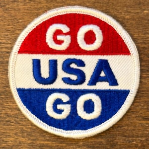 May include: A round patch with a red, white, and blue color scheme. The patch features the words "GO USA GO" in white, embroidered lettering. The patch has a white border and is likely an emblem or accessory.