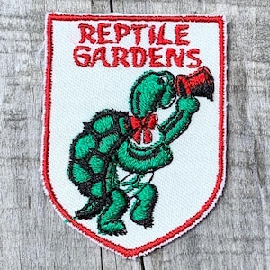 Op de afbeelding: Een schildvormige geborduurde patch met een witte achtergrond en een rode rand. Het toont een groene schildpad met een rode vlinderdas die een rode megafoon vasthoudt. De tekst REPTILE GARDENS is in rode letters bovenaan geborduurd.