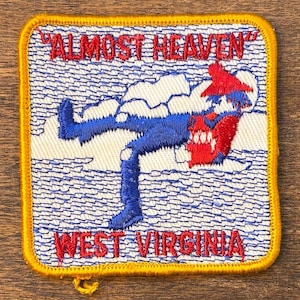 Op de afbeelding: Geborduurde patch met een gele rand. De patch toont de tekst "ALMOST HEAVEN" en "WEST VIRGINIA" in het rood. Het toont een cartoonfiguur in een blauwe broek en een rode hoed, die door wolken valt. De achtergrond is wit met blauwe lijnen.