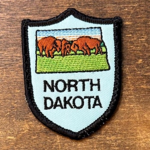 May include: A shield-shaped embroidered patch with a light blue background and a black border. The centre features a rectangular scene of two brown bison grazing on a green field under a blue sky. The text NORTH DAKOTA is stitched in black.