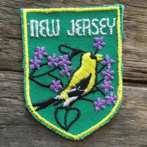 Puede incluir: Un parche bordado en forma de escudo con fondo verde y borde amarillo. Presenta un jilguero amarillo y negro posado sobre una rama con flores moradas. El texto NEW JERSEY está bordado en blanco en la parte superior del parche.