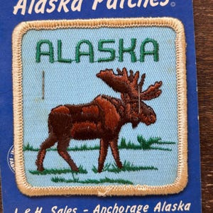 Puede incluir: Un parche bordado vintage que presenta un alce marrón sobre un fondo azul con la palabra "ALASKA" en letras verdes. El parche tiene un borde beige.