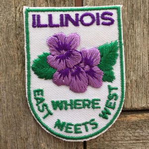 Peut inclure: Un écusson brodé blanc avec une bordure verte représentant des fleurs violettes au centre. Le texte ILLINOIS est brodé en violet en haut, et le texte WHERE EAST MEETS WEST est brodé en vert en bas de l'écusson.