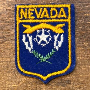 May include: A shield-shaped embroidered patch with a dark blue background and a yellow border. The word NEVADA is stitched in yellow at the top. Below, a yellow ribbon sits above a white star, white floral sprigs, and green crossed branches.