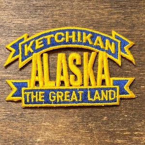 Könnte beinhalten: Gestickter Aufnäher mit dem Text "Ketchikan Alaska" in Gelb, mit blauem Banner und Rand. Der Schriftzug "THE GREAT LAND" befindet sich darunter. Der Aufnäher liegt auf einer Holzoberfläche.