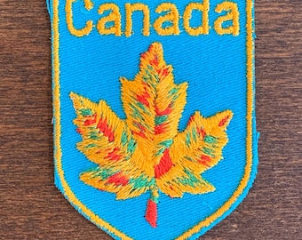 May include: A blue and yellow embroidered patch featuring a maple leaf and the word "Canada" in yellow.