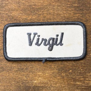 Könnte beinhalten: Ein rechteckiger gestickter Aufnäher mit weißem Stoffhintergrund und einem dicken, schwarz umstickten Rand. Der Name Virgil ist mittig in einer dunkelgrauen Schreibschrift eingestickt, was ein klares und gut lesbares Textdesign ergibt.