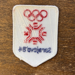 Puede incluir: Parche bordado blanco con los anillos ol&iacute;mpicos rojos y un emblema estilizado. El parche tiene el texto "Sarajevo 84" en azul. El parche es rectangular con un borde blanco y est&aacute; cubierto de pl&aacute;stico transparente.