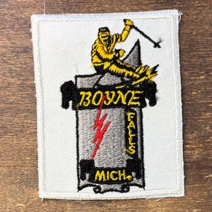 May include: A rectangular embroidered patch on a white background featuring a yellow skier in motion. The design includes a grey mountain shape, a red lightning bolt, and black text reading BOYNE FALLS MICH in a bold, stylized font.