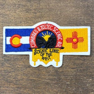 May include: Embroidered patch featuring the Colorado and New Mexico state flags, with text reading "Cumbres & Toltec Scenic RR" and "Scenic Line of the West." The patch has a white border and is likely for a jacket or bag.
