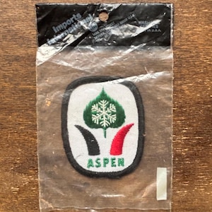 Puede incluir: Un parche bordado blanco, negro y rojo con la palabra "ASPEN" en letras negras. El parche presenta una hoja verde con un copo de nieve blanco en el centro. El parche está en una bolsa de plástico transparente con una tarjeta de encabezado negra que dice "Imports International".