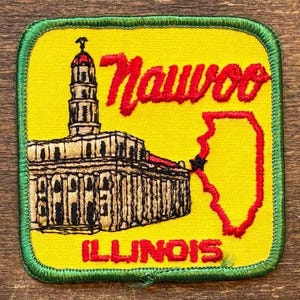 Op de afbeelding: Geborduurde patch met een gele achtergrond en groene rand. Het toont het woord "Nauvoo" in rood script, een gedetailleerde illustratie van een gebouw en de rode omtrek van Illinois. Het woord "Illinois" staat onderaan de patch.