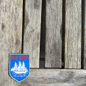 May include: A shield-shaped embroidered patch featuring a blue background with a white sailing ship graphic and the red text NEW JERSEY at the top. The patch has a yellow border and is designed for attachment to fabric items like clothing or bags.