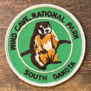 May include: A circular embroidered patch featuring a brown and tan prairie dog centered on a bright green background. The text Wind Cave National Park South Dakota is stitched in dark brown thread along the border with a white merrowed edge.