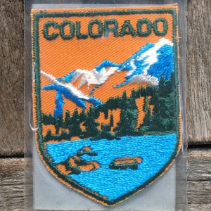 Könnte beinhalten: Ein vintage gestickter Aufnäher mit dem Wort "COLORADO" in grünen Buchstaben auf orangefarbenem Hintergrund. Der Aufnäher zeigt eine Bergkette mit schneebedeckten Gipfeln, einem See und Bäumen in Grün.