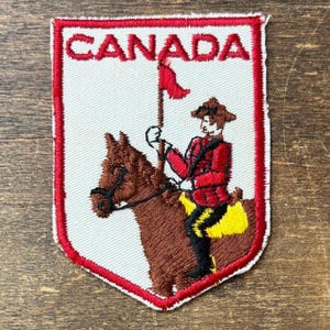 Könnte beinhalten: Ein schildförmiger, bestickter Aufnäher mit weißem Hintergrund und rotem Rand. Der Schriftzug CANADA ist oben in Rot eingestickt. Darunter reitet ein braunes Pferd mit einer Figur in roter Jacke, schwarzer Hose und braunem Hut, die eine rote Fahne hält.
