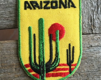 Puede incluir: Un parche bordado amarillo con la palabra "ARIZONA" en letras negras. El parche presenta una imagen estilizada de tres cactus saguaro con un sol rojo detrás de ellos.