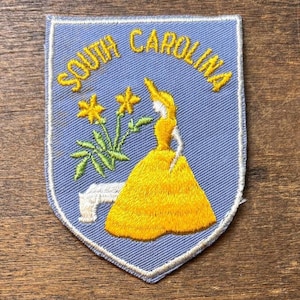 May include: Embroidered patch featuring the state of South Carolina. The patch has a light blue background with the words "SOUTH CAROLINA" in yellow. It depicts a woman in a yellow dress, yellow flowers, and a white bench.