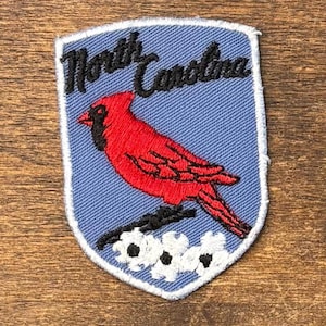 May include: A shield-shaped embroidered patch with a light blue background. It features a bright red cardinal bird perched on a branch above three white dogwood flowers. The text North Carolina is embroidered in black script at the top.