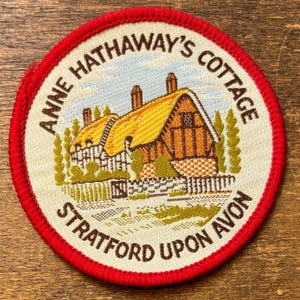May include: A round patch with a red border featuring an illustration of Anne Hathaway's Cottage in Stratford Upon Avon. The cottage has a yellow thatched roof and brown timber framing. The text "ANNE HATHAWAY'S COTTAGE" and "STRATFORD UPON AVON" are in brown.