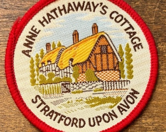 Può includere: Una toppa rotonda con bordo rosso raffigurante il cottage di Anne Hathaway a Stratford Upon Avon. Il cottage ha un tetto di paglia giallo e struttura in legno marrone. Il testo "ANNE HATHAWAY'S COTTAGE" e "STRATFORD UPON AVON" è marrone.