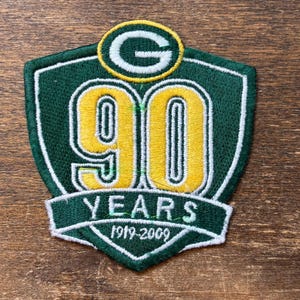 May include: A green and yellow embroidered patch commemorating 90 years. The patch features a shield shape with the number "90" in yellow, the text "YEARS" and the years "1919-2009". The top of the patch has a green "G" logo.