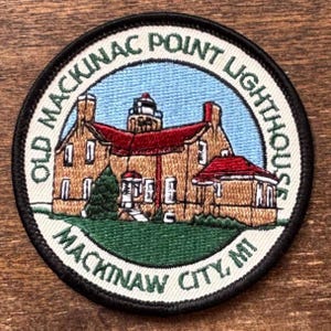 Könnte beinhalten: Runder, gestickter Aufnäher mit dem Old Mackinac Point Lighthouse in Mackinaw City, MI. Das Design zeigt das Leuchtturmgebäude mit rotem Dach und grünem Gras. Der Text "OLD MACKINAC POINT LIGHTHOUSE" und "MACKINAW CITY, MI" ist kreisförmig um das Bild angeordnet.