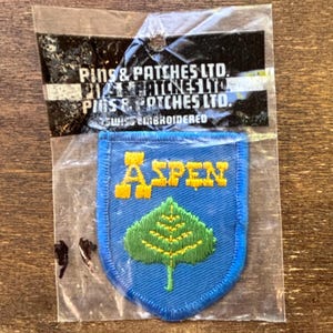 Op de afbeelding: Een blauwe en gele geborduurde patch met het woord "ASPEN" in gele letters en een gestileerd groen espenblad op een blauwe achtergrond. De patch is verpakt in een doorzichtige plastic zak met een wit label waarop staat "Pins & Patches Ltd. Swiss Embroidered".
