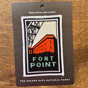 Könnte beinhalten: Ein rechteckiger Aufnäher mit einem stilisierten Bild von Fort Point, mit einer Brückenstruktur und einem roten Gebäude. Der Text "FORT POINT" ist grün, und der Aufnäher ist weiß umrandet. Der Text "THE GOLDEN GATE NATIONAL PARKS" steht unten.