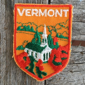 May include: Embroidered patch featuring the word "VERMONT" in white, above a church and landscape scene. The patch has an orange background with green and red accents, and a red border. The patch is shield-shaped.