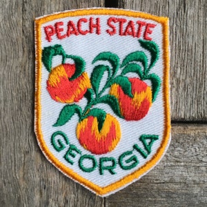 May include: Embroidered patch featuring a shield design with the words "PEACH STATE GEORGIA". The patch showcases three peaches in shades of red and orange with green leaves, outlined in yellow and white.