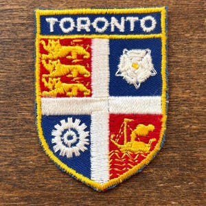 Puede incluir: Un parche bordado en forma de escudo con la palabra TORONTO en letras blancas sobre un fondo azul. El escudo está dividido en cuatro cuadrantes con una cruz blanca, mostrando tres leones amarillos, una rosa blanca, un engranaje blanco y un barco amarillo.