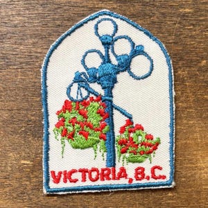 May include: Embroidered patch featuring a blue lamppost with hanging baskets of red flowers and green foliage. The patch has a white background and a blue border. The text "VICTORIA, B.C." is displayed in red.