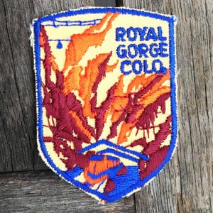 May include: A shield-shaped embroidered patch with a light yellow background. It features a blue border and blue text reading ROYAL GORGE COLO. The design depicts orange and maroon flames rising above a blue bridge structure and water.