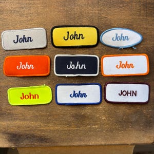 May include: Nine embroidered patches displaying the name "John". The patches are in various shapes and colours, including grey, yellow, orange, navy, white, and green. The patches have different border colours and fonts. The patches are rectangular and oval.