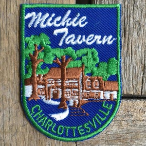 May include: A rectangular embroidered patch with a blue background and a green border. It features a brown building surrounded by green trees and white accents. The text "Michie Tavern" is in white, and "CHARLOTTESVILLE" is in green at the bottom.