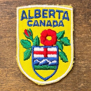 Könnte beinhalten: Ein schildförmiger, bestickter Aufnäher mit gelbem Hintergrund. Der Text ALBERTA CANADA ist oben in Blau gestickt. Unter dem Text befindet sich eine rote Wildrose mit grünen Blättern und ein Wappenschild mit einem roten Kreuz, blauem Himmel und grünen Hügeln.