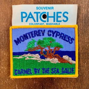 May include: A blue and yellow embroidered patch with the text "Monterey Cypress Carmel by the Sea Calif." and a depiction of a cypress tree. The patch is labeled "Souvenir Patches Colorfast, Washable."