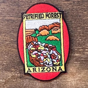Puede incluir: Parche bordado con las palabras "Petrified Forest Arizona" en un diseño rectangular, dentro de un óvalo rojo. El diseño incluye un paisaje con formaciones rocosas coloridas y hierba verde.