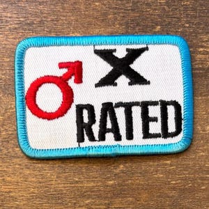 May include: A rectangular embroidered patch with a white background and a bright blue border. It features a red male gender symbol, a large black letter X, and the word RATED in black capital letters, all stitched onto the white fabric surface.