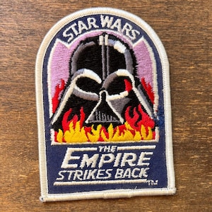 May include: An embroidered patch featuring a black Darth Vader helmet graphic centered against a light purple background with red and yellow flames at the base. The text STAR WARS appears at the top, and THE EMPIRE STRIKES BACK is at the bottom.