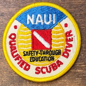 Pode incluir: Um emblema bordado circular com uma seção superior azul com o texto branco NAUI. O centro exibe um quadrado vermelho com uma faixa diagonal branca, cercado por um sol amarelo. A borda branca diz QUALIFIED SCUBA DIVER.